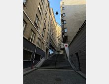 Тясна градска улица между високи жилищни сгради, водеща към широко стълбище с метални парапети. Над улицата са опънати кабели с висящи улични лампи, а вдясно има знак „влизането забранено“.