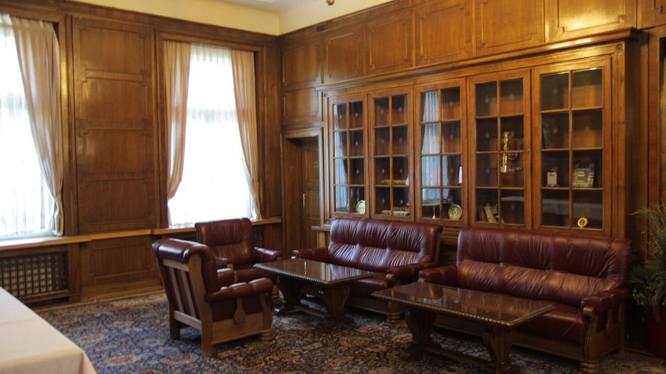 Класически салон с дървена ламперия по стените, високи прозорци с бели завеси и полилей на тавана. Вътре има кожени дивани и кресла около ниски маси върху голям орнаментиран килим, а по стената има вграденa остъкленa библиотека/витрина.