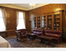 Класически салон с дървена ламперия по стените, високи прозорци с бели завеси и полилей на тавана. Вътре има кожени дивани и кресла около ниски маси върху голям орнаментиран килим, а по стената има вграденa остъкленa библиотека/витрина.