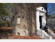 Стара мазилка с малък прозорец на ниска сграда, почти изцяло обрасла с голи увивни растения, с бял входен портал и стълби към тъмна врата. Около сградата има дървета и паднали есенни листа на земята.