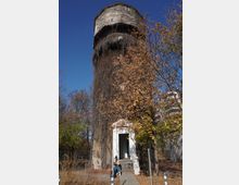 Висока цилиндрична водна кула с многоъгълна надстройка и малки прозорци, обрасла с бръшлян и сухи лиани. В основата има вход с бетонни стъпала, заобиколен от есенни дървета и ограда под ясно синьо небе.