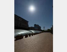 Широка градска пешеходна алея с настилка от павета и редица саксии, до която се простират ниски полукръгли стъклени куполи като покрив на подземно пространство. В далечината се виждат високи административни сгради и монументална колона на площад, под силно слънце в ясно небе.