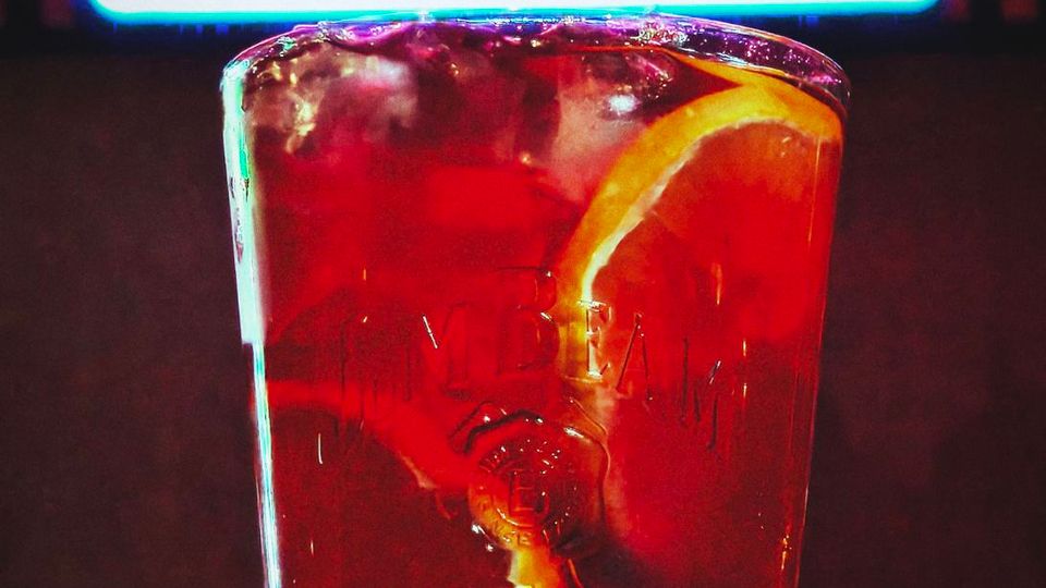 Неонова табела с надпис „BAR“ на тъмен фон, заснета в близък план. На преден план има висока чаша с лед и напитка, гарнирана с резен портокал.