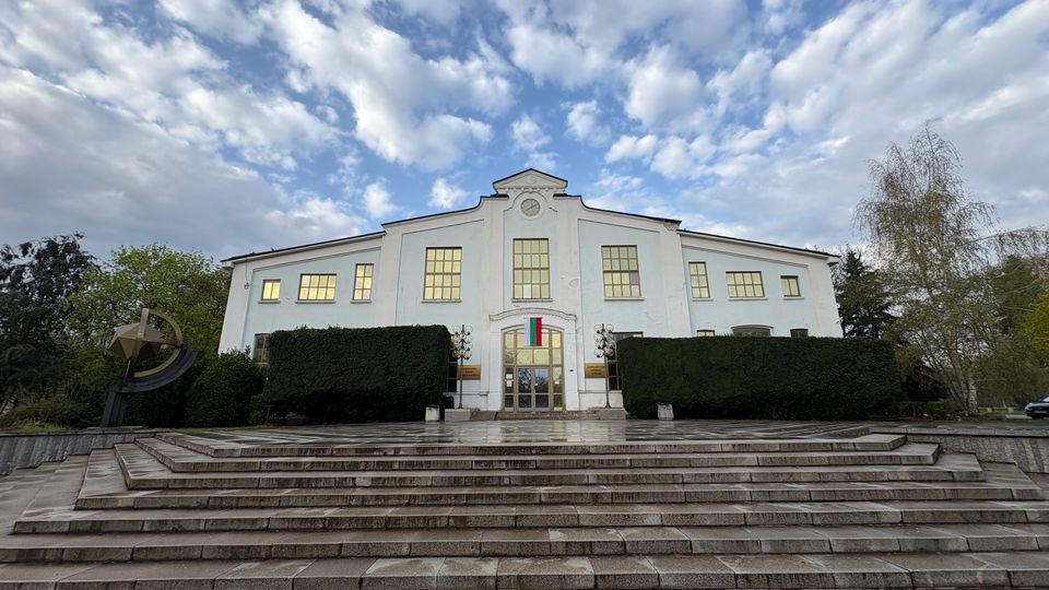 Фасада на голяма бяла обществена сграда с централно разположен вход, широки прозорци и часовник над него. Отпред има широки каменни стълби, подрязани живи плетове и дървета от двете страни под облачно небе.