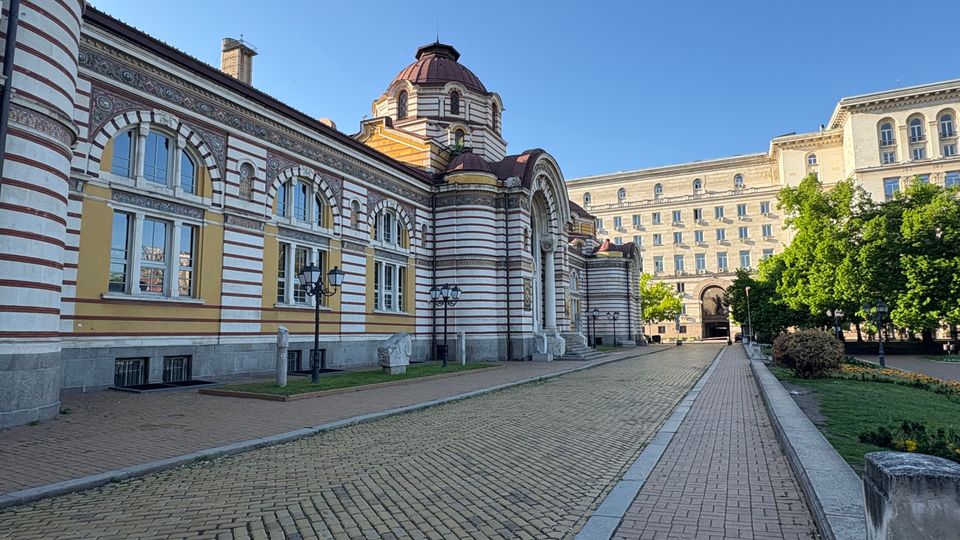 Изглед към голяма историческа сграда с фасада на червено-бели хоризонтални ивици, аркови прозорци и централен купол над вход с висока арка. Пред нея има павирана алея с улични фенери, тревни площи и дървета, а в далечината се вижда висок правоъгълен административен корпус.