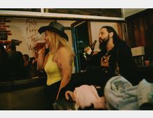 Интериор на малък бар с дървени греди и облицовка, отворена входна врата и барплот с бутилки и чаши. На стената има графит „Pink Floyd The Wall“ и частична тухлена декорация.