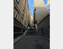 Тясна градска уличка с калдъръм, която води към широки стълби с парапети, разположени между високи жилищни сгради с прозорци и балкони. Над стълбите има опънати кабели и висящи улични лампи, а отгоре се вижда открито синьо небе.