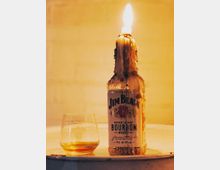 Бутилка бърбън върху маса, покрита с разтекъл се восък и запалена като свещ, с пламък на гърлото. До нея има ниска чаша с малко кехлибарена напитка на неутрален светъл фон.