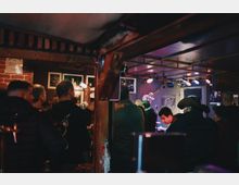 Интериор на малък музикален бар с тухлена стена, дървени греди и рамкирани снимки по стените. В дъното има ниска сцена с микрофон и сценично осветление, а над нея се виждат плакати и декоративна китара.