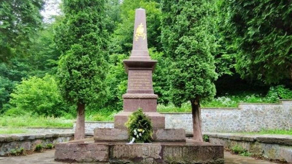 A stone monument featuring a tall, tapered obelisk engraved with gold text, flanked by two tall evergreen trees. The monument is set on a raised stone platform surrounded by greenery, with a low stone wall partly visible in the background.
