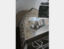 Гледка отгоре към тясно спираловидно стълбище с бетонни стъпала и бели метални парапети, спускащо се през няколко нива. Стълбищната клетка е с бели стени и малка площадка на дъното.