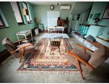 Интериор на малка всекидневна или общо помещение с зелени стени, дълъг прозоречен ред и килим в центъра с ниска маса и два ретро фотьойла. В дъното има бяла дъска и дървен шкаф, а вдясно — повдигната платформа със стъпала с шарени плочки, бар плот и вградени места за сядане с възглавници.