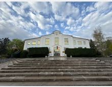 Фасада на голяма бяла обществена сграда с централно разположен вход, широки прозорци и часовник над него. Отпред има широки каменни стълби, подрязани живи плетове и дървета от двете страни под облачно небе.