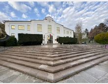 Бяла обществена сграда с симетрична фасада, големи правоъгълни прозорци и централен вход под фронтон с часовник и българско знаме. Пред нея има широка гранитна стълба и площадка, оградени от подрязани живи плетове и дървета.