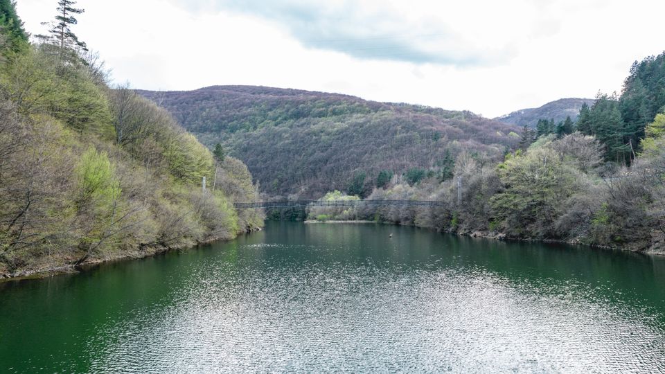 Широко езеро или язовир, заобиколено от гористи хълмове, с зелена вода и леко вълнение на повърхността. В далечината се вижда тесен мост, пресичащ водата, под облачно небе.