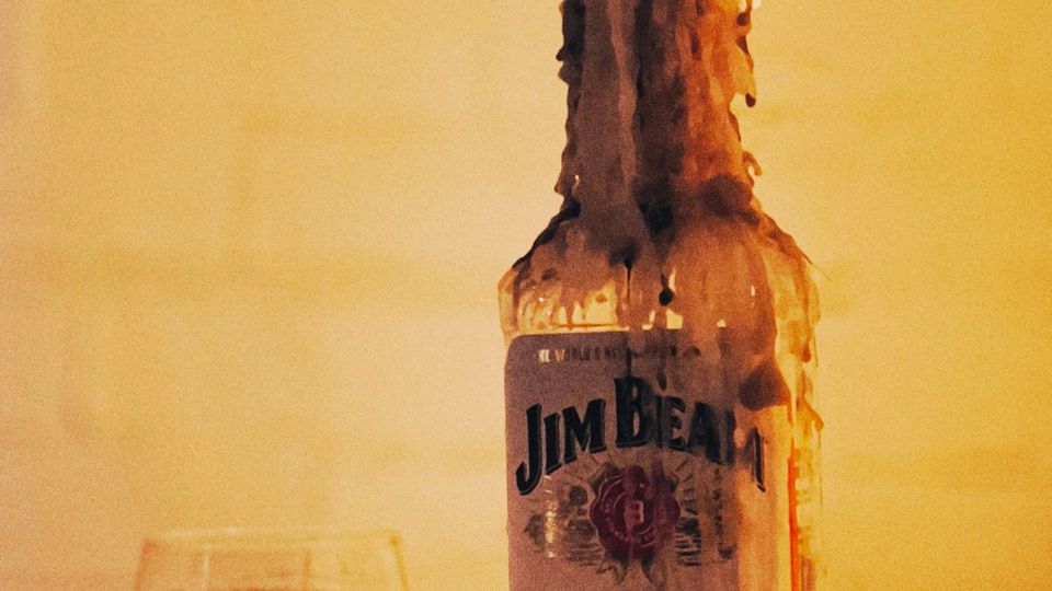 Бутилка бърбън върху маса, покрита с разтекъл се восък и запалена като свещ, с пламък на гърлото. До нея има ниска чаша с малко кехлибарена напитка на неутрален светъл фон.