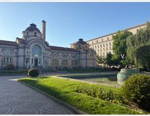 Историческа обществена сграда с куполи и декоративна фасада, разположена до малък парк с алеи, тревни площи и правоъгълно водно огледало с фонтан. На заден план се вижда висока административна сграда и дървета около водата.