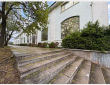 Дълга светла сграда с големи арковидни прозорци и висока фасада, заснета отстрани покрай широки бетонни стъпала. Отпред има ниски храсти и дървета край алея, водеща по протежение на сградата.