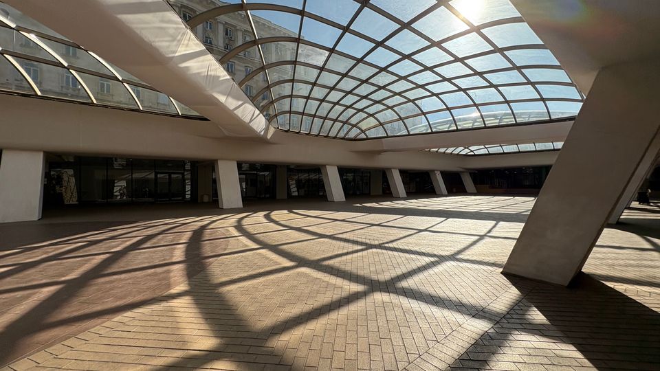 Покрит вътрешен двор в модерна сграда с голямо извито стъклено покритие на метална решетка, което пропуска слънчева светлина и хвърля геометрични сенки върху настилка от тухлени павета. Пространството е обградено от наклонени колони и редица остъклени входове по периметъра.