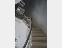 Вътрешно стълбище в сграда с каменни стъпала и извита дървена ръкохватка върху бяла метална парапетна решетка. Стълбите се изкачват покрай високи светлосиви стени към горно ниво с декоративни елементи по тавана.