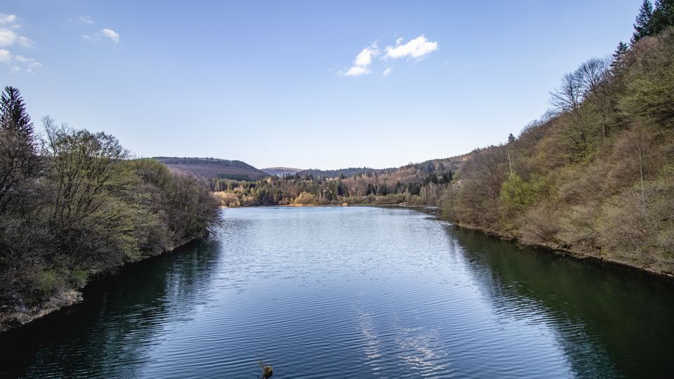 Спокойно езеро, разположено в долина между ниски гористи хълмове, с дървета по двата бряга и отражения във водата. На заден план се виждат далечни възвишения под ясно синьо небе с единични облаци.