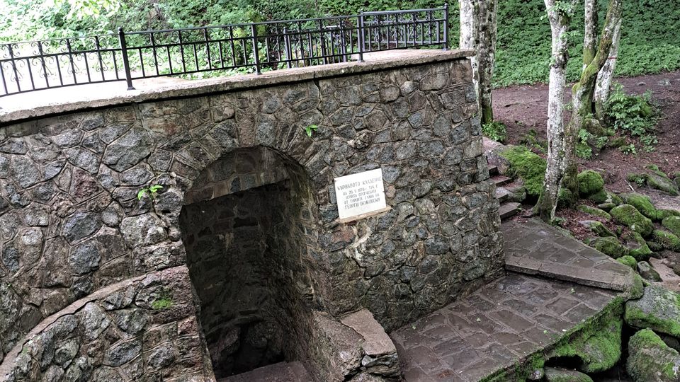 A stone structure with an arched entrance sits next to a small stream flowing over moss-covered rocks in a lush forest setting. The area is surrounded by trees and features a metal railing along the top of the structure for safety.