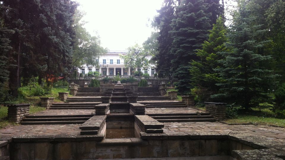 Каскадна каменна тераса с басейн и стъпаловидни платформи, водещи към бяла класическа сграда с колонада на заден план. Мястото е обградено от високи иглолистни дървета и зеленина в парк.