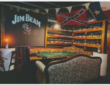 Интериор на бар с дървени рафтове по стените, плътно подредени с бутилки алкохол, и голям надпис „Jim Beam“ на тъмна стена. В центъра има маса за покер със зелено сукно и винтидж мека мебел, а над рафтовете висят знамена и декоративни гирлянди.