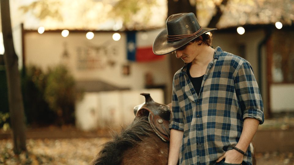 Открит селски двор или ранчо с дървета и наниз от лампички, на заден план се вижда ниска постройка със знаме на Тексас. На преден план има оседлан кон, а земята е покрита с паднали листа.