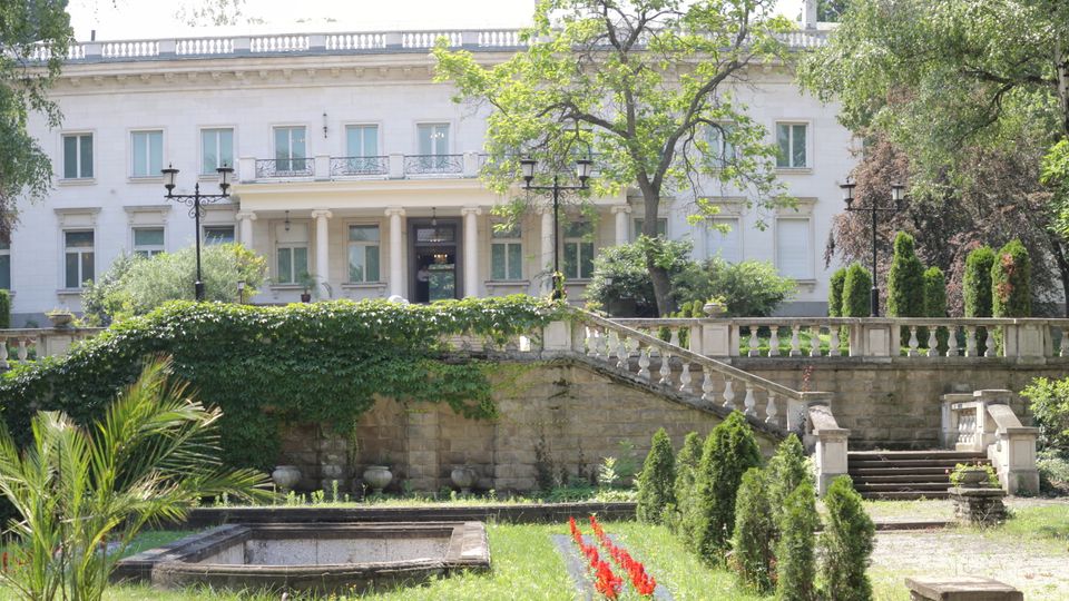 Бяла двуетажна неокласическа сграда с колонада и балкон, разположена зад терасирана градина. На преден план има каменни стълби и парапет, обрасли с бръшлян, алеи и цветни лехи сред високи дървета.