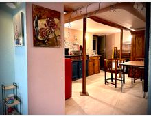 Интериор на малка кухня и трапезария с нисък таван и видими греди, осветление и гирлянди. Виждат се дървени шкафове с мозайка зад мивката, червен хладилник, маса със столове и голям дървен гардероб.