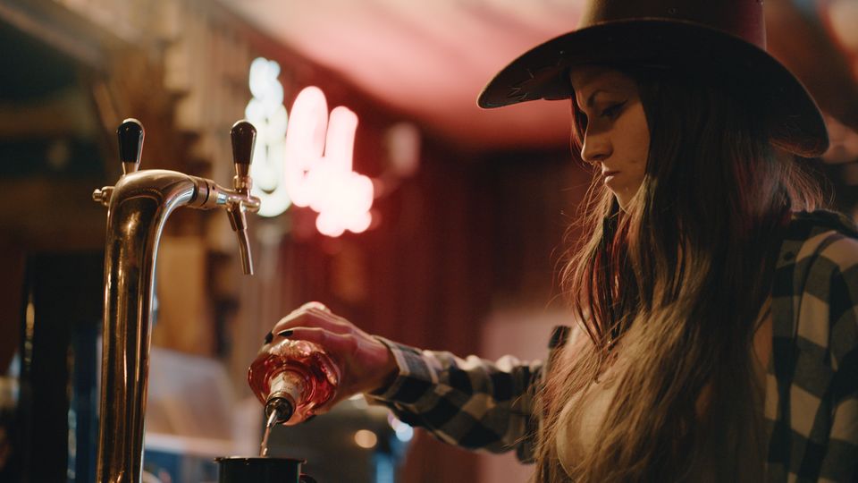 Интериор на бар с дървен плот и метална бирена колона с кранове на преден план. В дъното се вижда размазан неонов надпис и топло, приглушено осветление.