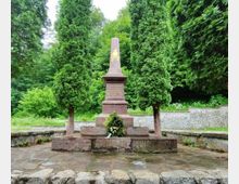 A stone monument featuring a tall, tapered obelisk engraved with gold text, flanked by two tall evergreen trees. The monument is set on a raised stone platform surrounded by greenery, with a low stone wall partly visible in the background.
