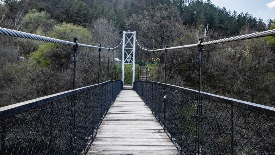 Висящ пешеходен мост с дървена настилка и метални мрежести парапети, водещ към бяла метална конструкция в средата. Мостът преминава над гориста долина с хълмове на заден план под частично облачно небе.