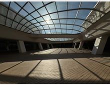 Широко вътрешно атриумно пространство с извит стъклен покрив в метална решетка, през който навлиза слънчева светлина. Подът е с тухлени плочи, а бели наклонени колони и сенките от покрива оформят геометрични линии по настилката.