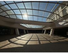 Покрит вътрешен двор или пасаж с голям извит стъклен покрив на метална решетка, през който влиза силна дневна светлина. На пода от павета се виждат решетъчни сенки, а пространството е обградено от масивни колони и фасади на сгради отстрани.