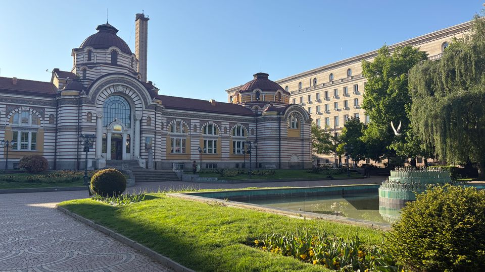 Историческа обществена сграда с куполи и декоративна фасада, разположена до малък парк с алеи, тревни площи и правоъгълно водно огледало с фонтан. На заден план се вижда висока административна сграда и дървета около водата.