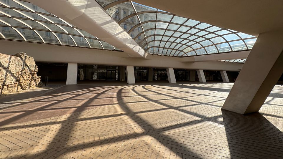 Покрито атриумно пространство с извита стъклена покривна конструкция и масивни наклонени колони, над настилка от тухлени павета. Вляво има открити каменни руини, а в дъното се вижда редица стъклени входове и фасада на сгради през покрива.