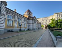 Изглед към голяма историческа сграда с фасада на червено-бели хоризонтални ивици, аркови прозорци и централен купол над вход с висока арка. Пред нея има павирана алея с улични фенери, тревни площи и дървета, а в далечината се вижда висок правоъгълен административен корпус.