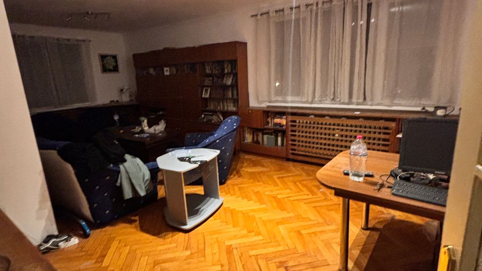 Interior living room with herringbone parquet wood flooring, a blue sofa and armchair around a small coffee table, and a tall bookcase along the back wall. Large windows with sheer curtains line the right side above a radiator cover, with a simple ceiling track light and a small desk near the doorway.