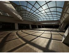 Широко вътрешно атриумно пространство в модерна сграда с извита стъклена покривна конструкция на метална решетка. Слънчевата светлина през покрива хвърля мрежести сенки върху тухлената настилка и белите наклонени колони около периметъра.