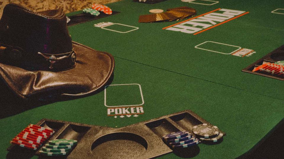 Зелена маса за покер в казино с обозначени места за играчи, отделения за чипове и централен отвор за събиране. По ръба са подредени купчини покер чипове и се виждат подплатени кожени подлакътници.