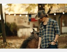 Открит селски двор или ранчо с дървета и наниз от лампички, на заден план се вижда ниска постройка със знаме на Тексас. На преден план има оседлан кон, а земята е покрита с паднали листа.