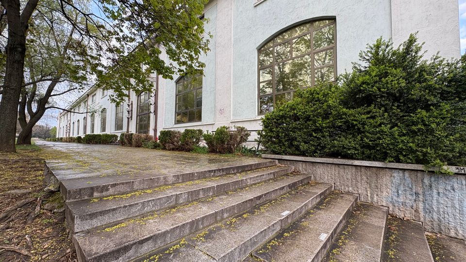 Дълга светла сграда с големи арковидни прозорци и висока фасада, заснета отстрани покрай широки бетонни стъпала. Отпред има ниски храсти и дървета край алея, водеща по протежение на сградата.