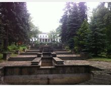 Каскадна каменна тераса с басейн и стъпаловидни платформи, водещи към бяла класическа сграда с колонада на заден план. Мястото е обградено от високи иглолистни дървета и зеленина в парк.