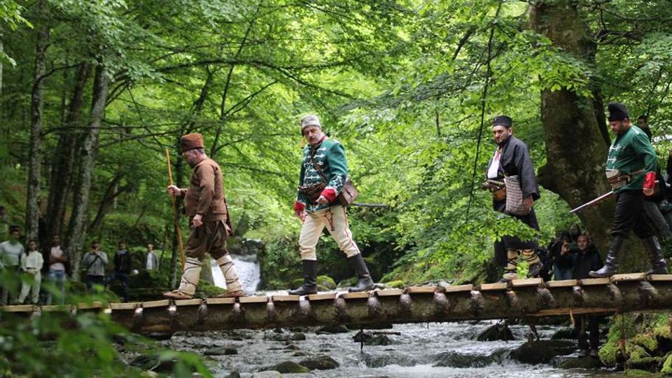 A wooden footbridge spans a shallow, rocky stream in a dense forest of tall green trees. The area is shaded and surrounded by lush vegetation, with small groups of people visible in the background.