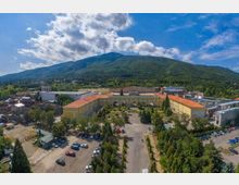 Въздушен изглед към голям комплекс от ниски сгради с жълти фасади и червени керемидени покриви, оформящи вътрешен двор с алеи, зелени площи и паркинг. На заден план се вижда горист планински склон под частично облачно небе.