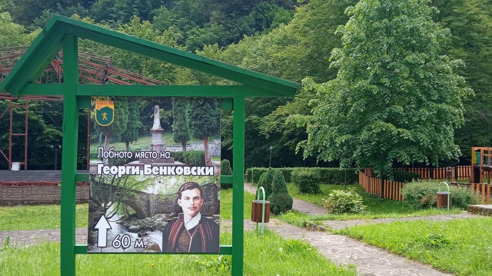 A small park with a wooden sign featuring an image and text in Cyrillic, set against a backdrop of dense, green forest. The area includes paved walkways, neatly maintained grass, and trees, with a wooden fence and playground elements visible in the distance.