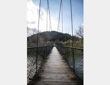 Дървен пешеходен висящ мост с метални въжета и мрежести парапети, простиращ се над река към нисък горист хълм. От двете страни има дървета и храсти, а над тях се вижда светло небе с облаци.