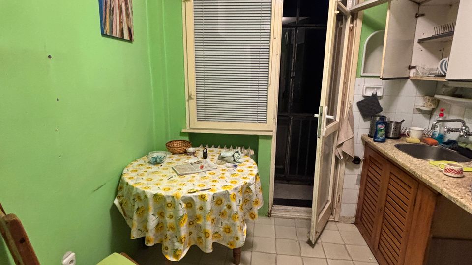 Small kitchen with green painted walls, a tiled floor, and a round table with a patterned tablecloth beside a window with blinds. To the right is a countertop with sink and white upper cabinets, and an open door leading to a dark balcony or exterior space.